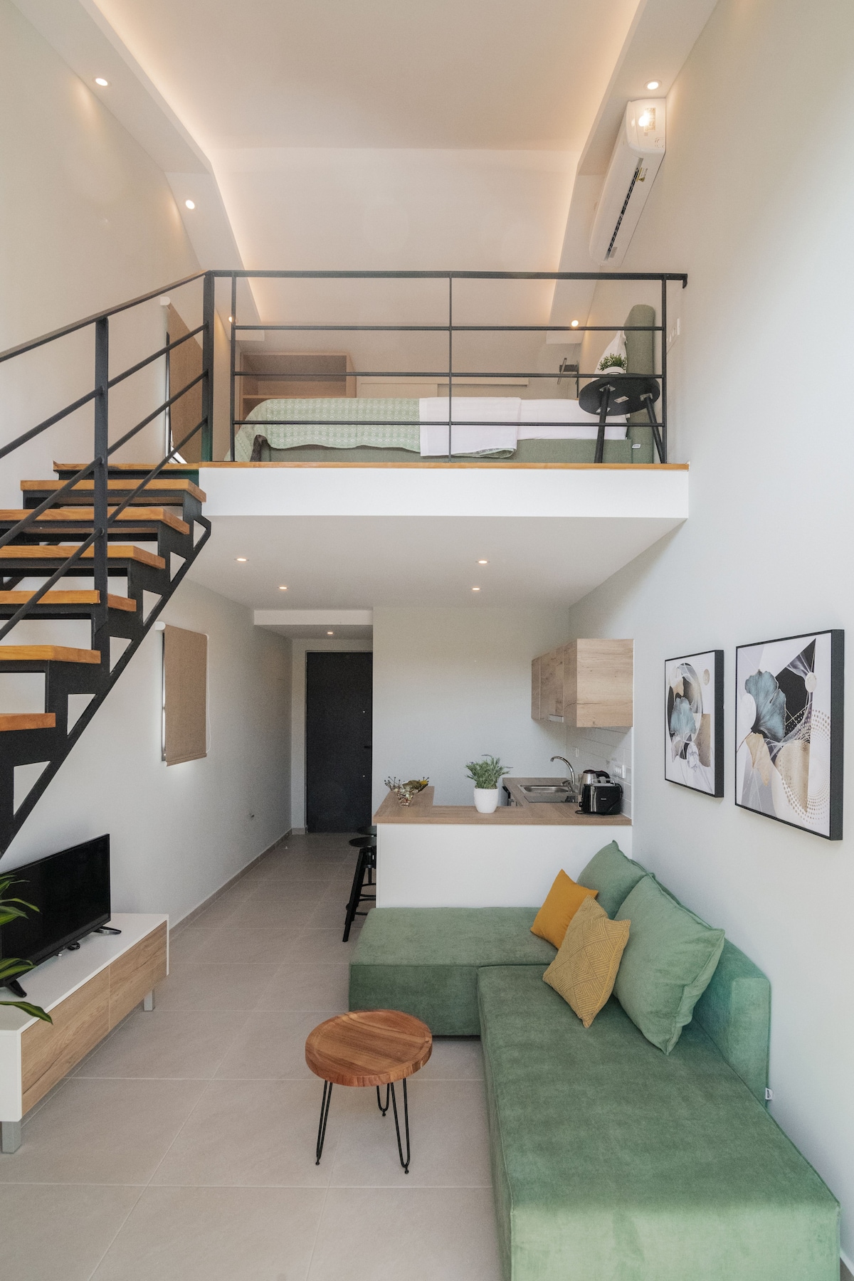 An open-plan layout reveals a cozy living area featuring a green sofa and a wall-mounted TV. A staircase leads to the upper level, where a comfortable bedroom is visible. Natural light fills the space, enhancing the modern interior decor.