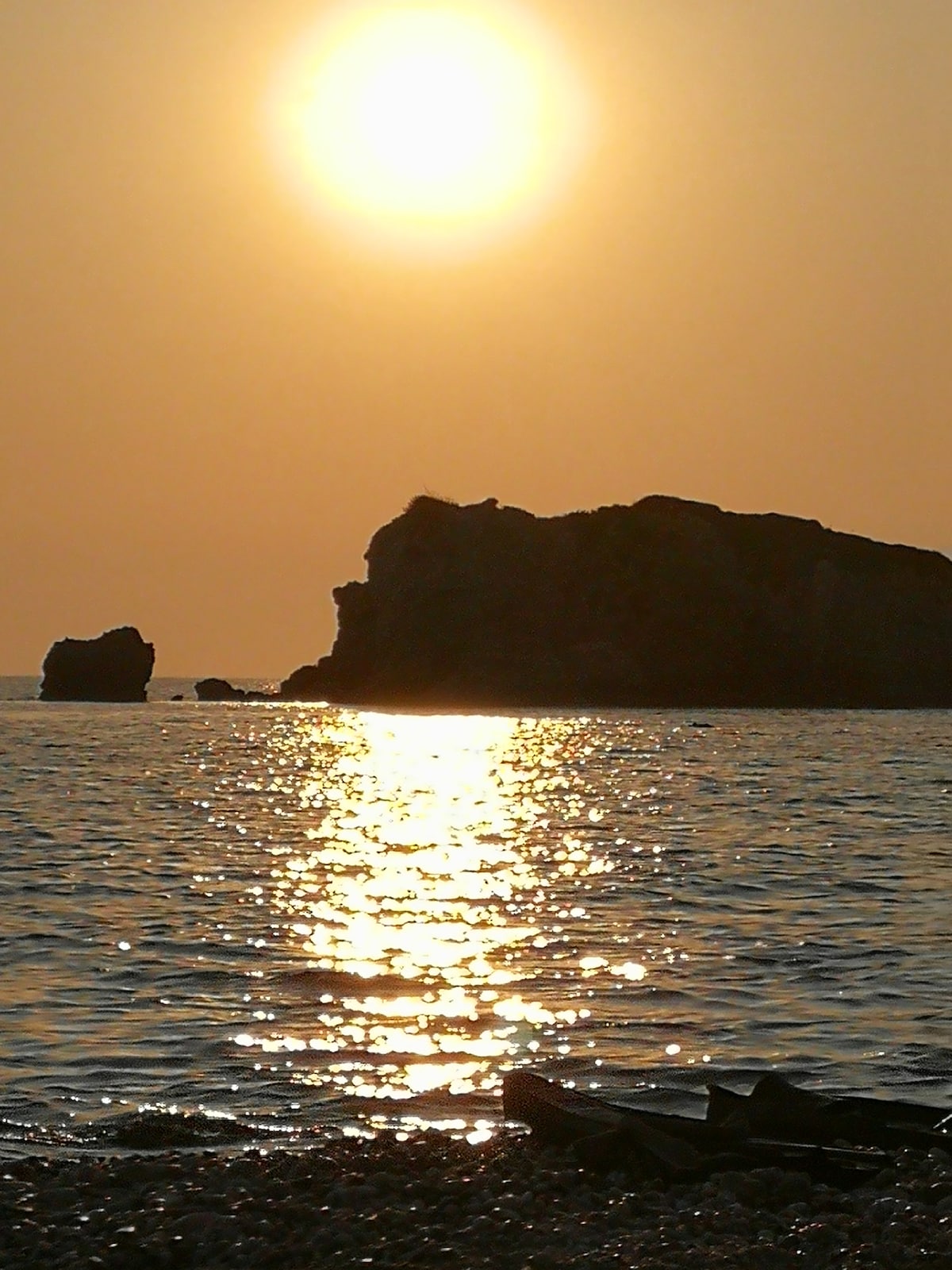 A tranquil sunset scene is captured over a calm sea, with the sun illuminating the horizon. Reflections of golden light shimmer on the water's surface, while a dark rock formation is positioned against the orange sky.