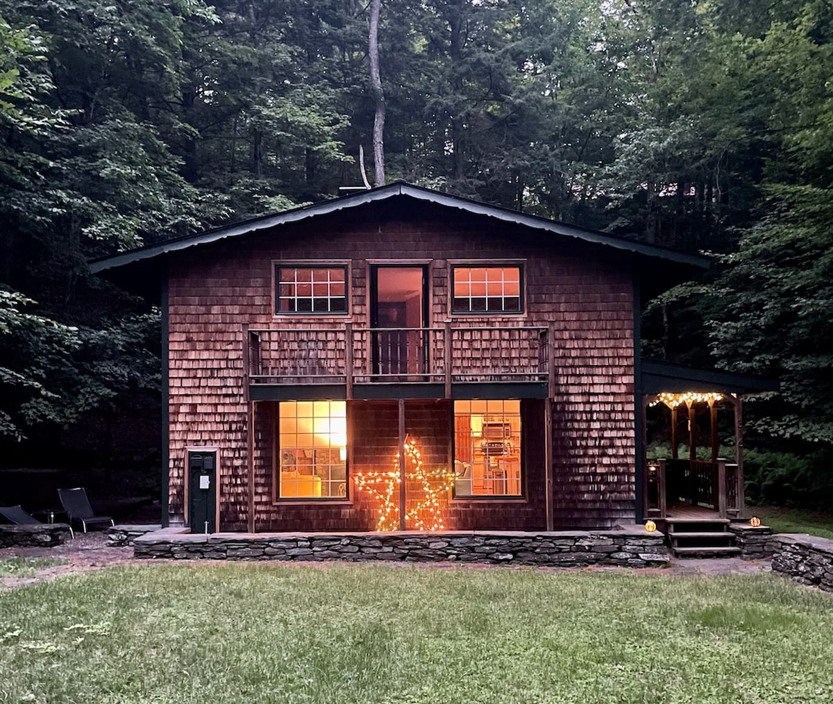 The exterior of a classic Vermont ski house is visible, featuring a wooden facade with a cozy, rustic charm. Large windows illuminate the main floor, where warm light glows. A star-shaped light decorates the front lawn, surrounded by a grassy area and bordered by trees.