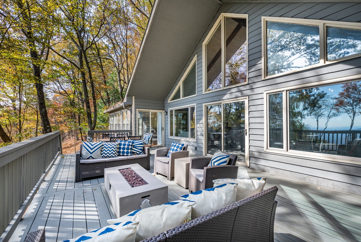 A spacious outdoor deck is surrounded by trees with autumn foliage. Comfortable seating arrangements are provided with plush cushions, and a modern fire pit is positioned in the center. Large glass windows reflect the scenic view of the surrounding nature.