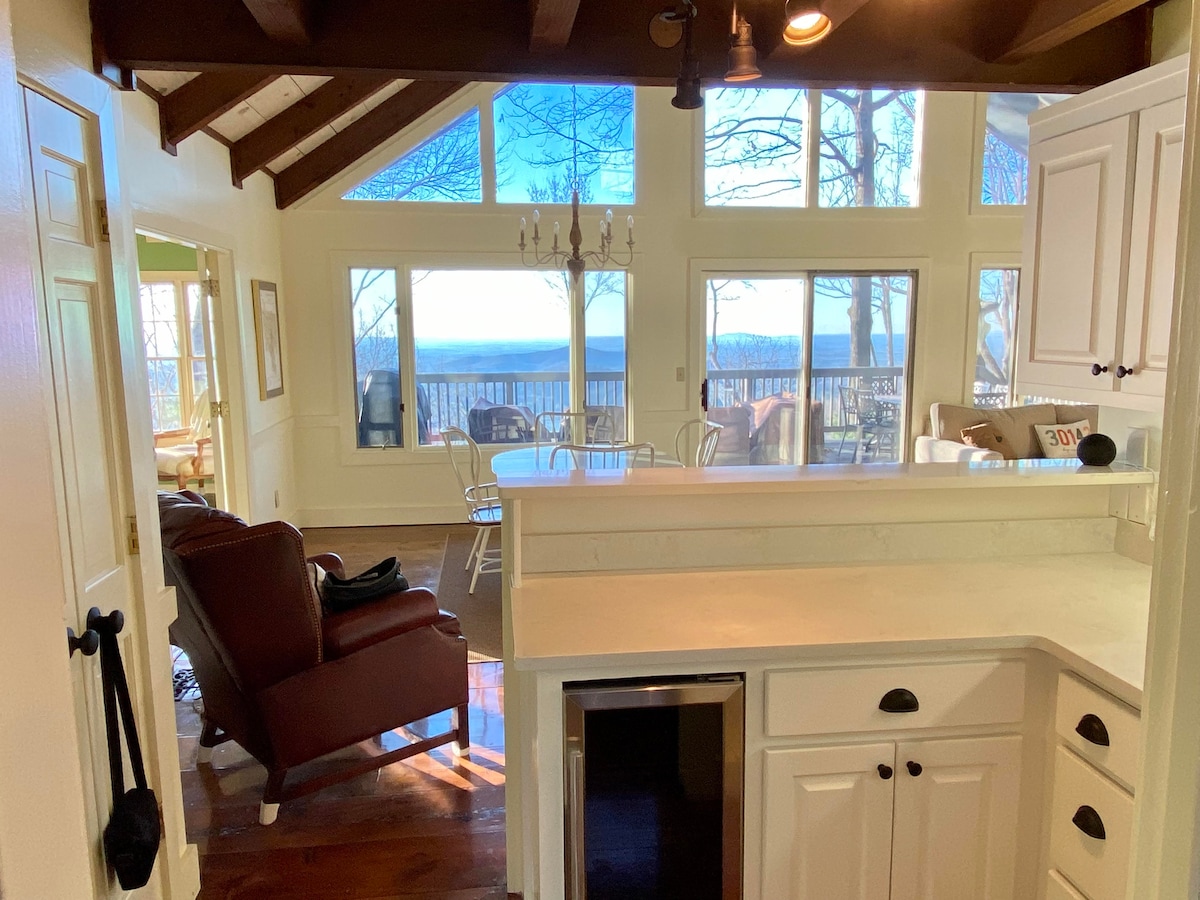 The open-concept living area features vaulted ceilings and large windows that frame views of the lake and surrounding nature. A dining table is positioned near the window, while comfortable seating is arranged in the cozy living space, inviting relaxation.