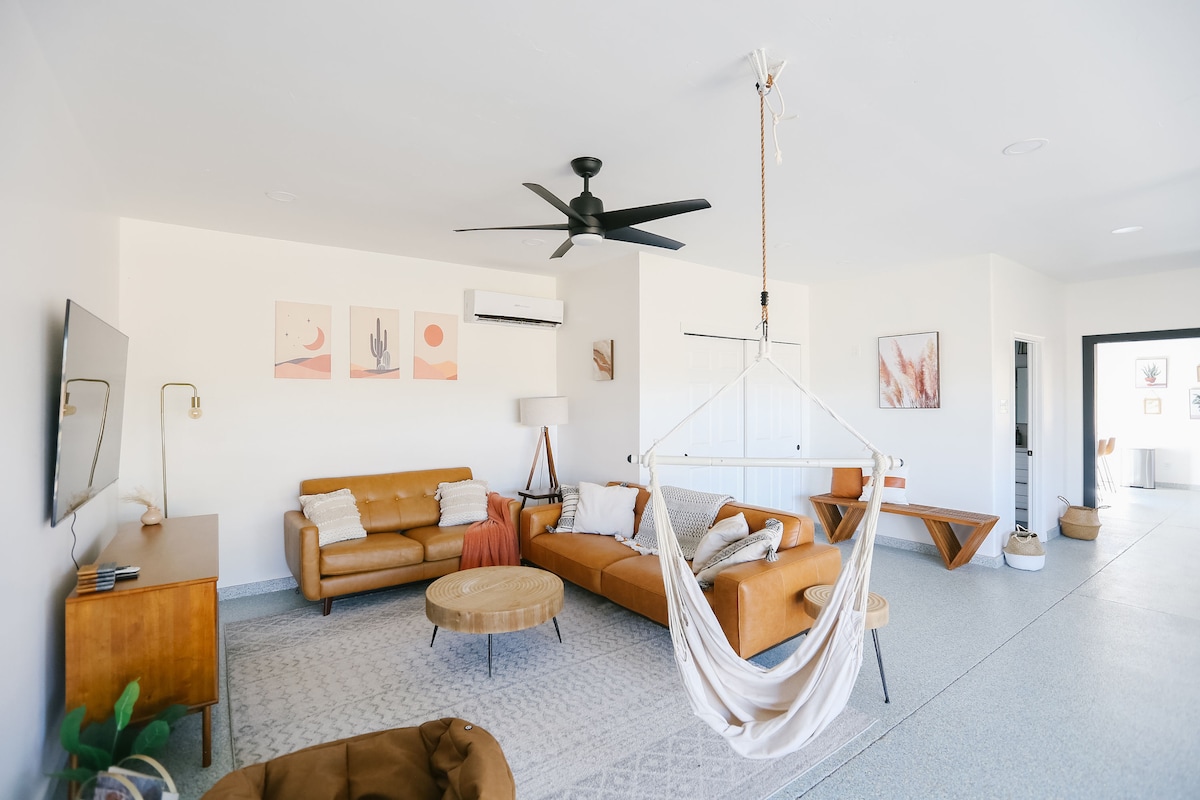 A spacious living area features two leather sofas and a round coffee table, complemented by decorative wall art. A ceiling fan provides airflow, and a hammock is suspended near the corner. Natural light illuminates the open layout, enhancing the inviting environment.
