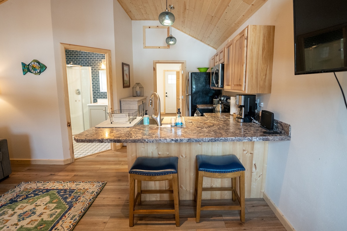 A welcoming kitchen space is equipped with stainless steel appliances and a large countertop with bar seating for two. Light wood cabinetry complements the open layout, while natural light enters through the adjacent living area. A doorway leads to a bathroom visible in the background.