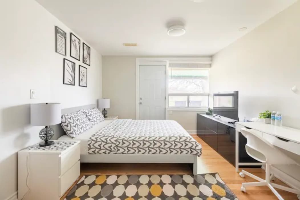 A bright bedroom features a spacious bed with a patterned blanket, complemented by artistic wall decor. A television is positioned on a sleek black dresser, and a white desk is provided for workspace, illuminated by natural light from the window.