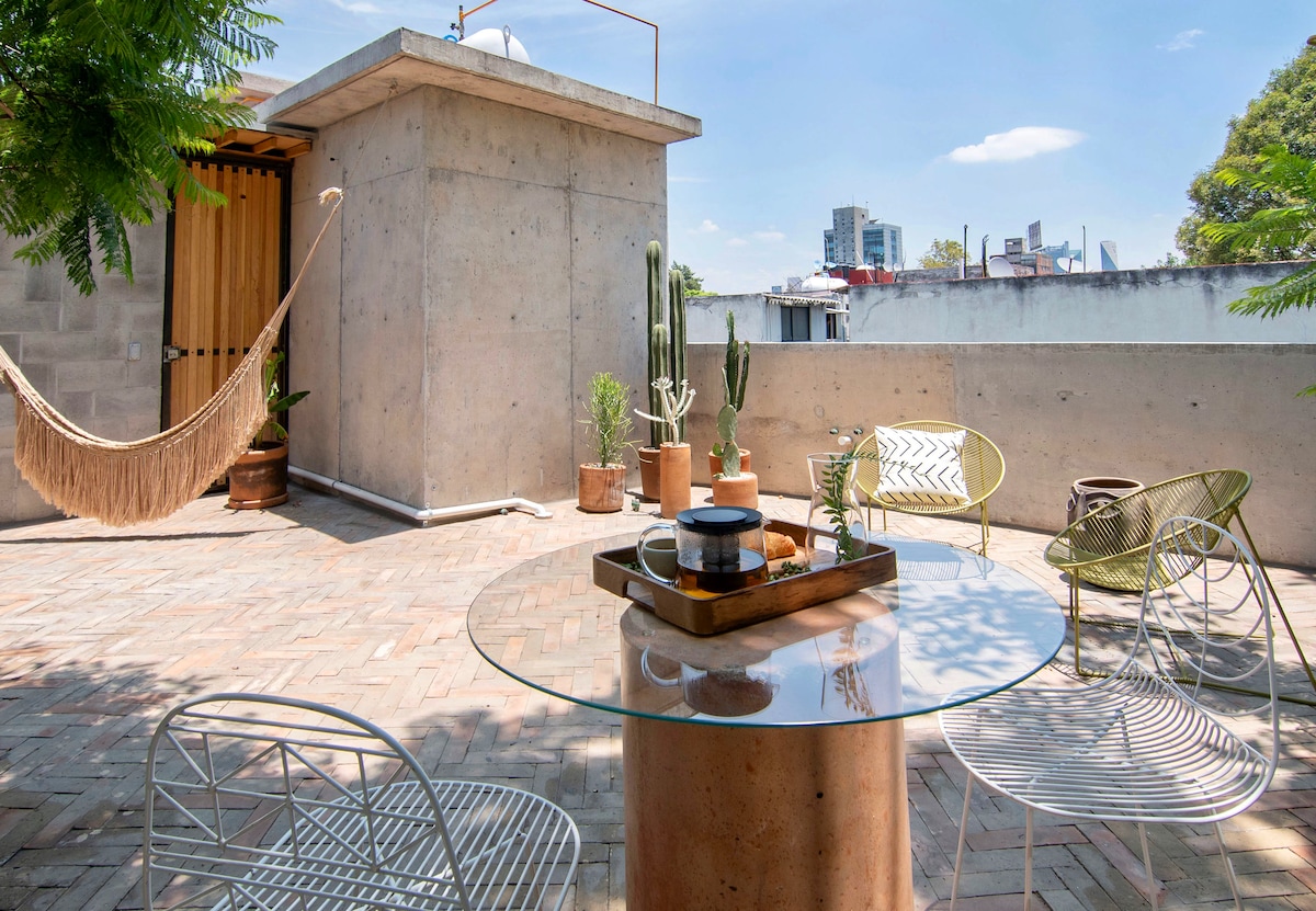An outdoor terrace features a round glass table atop a cylindrical concrete base, surrounded by metal chairs. Potted plants and cacti are positioned along the perimeter, and a hammock is gently hung to one side, all set against a clear blue sky.