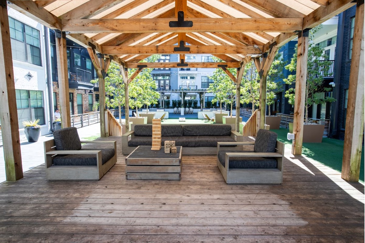 An open-air pavilion showcases comfortable seating arrangements with dark cushions on wooden frames. A central wooden table sits between the seating areas. Surrounding greenery is visible in the courtyard, creating a serene atmosphere for relaxation.