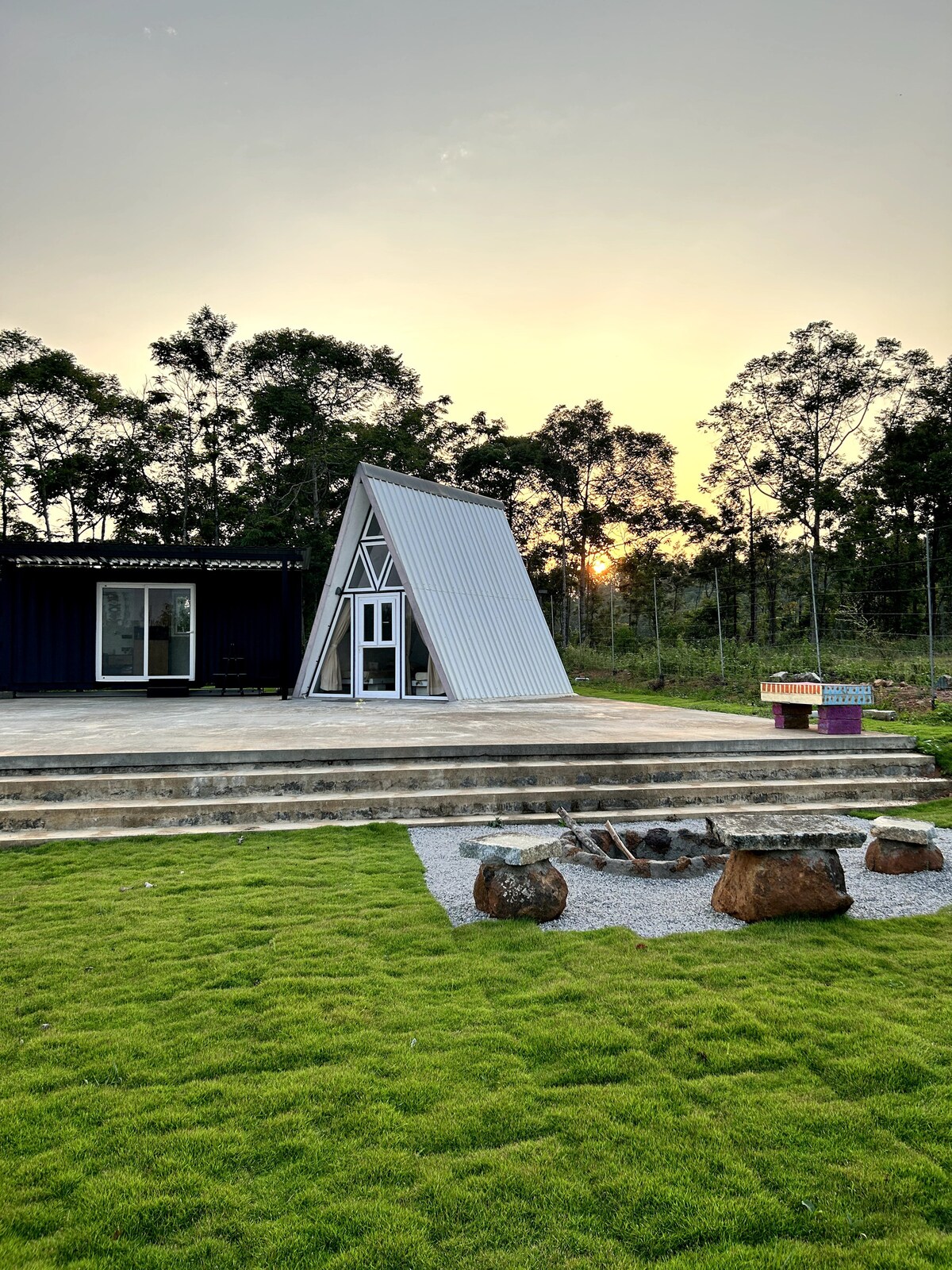 An A-frame cabin is set against a backdrop of greenery, with a gently sloped roof reflecting the soft hues of sunset. An outdoor patio of stone steps leads to a small fire pit area surrounded by smooth rocks, enhancing the serene outdoor space.