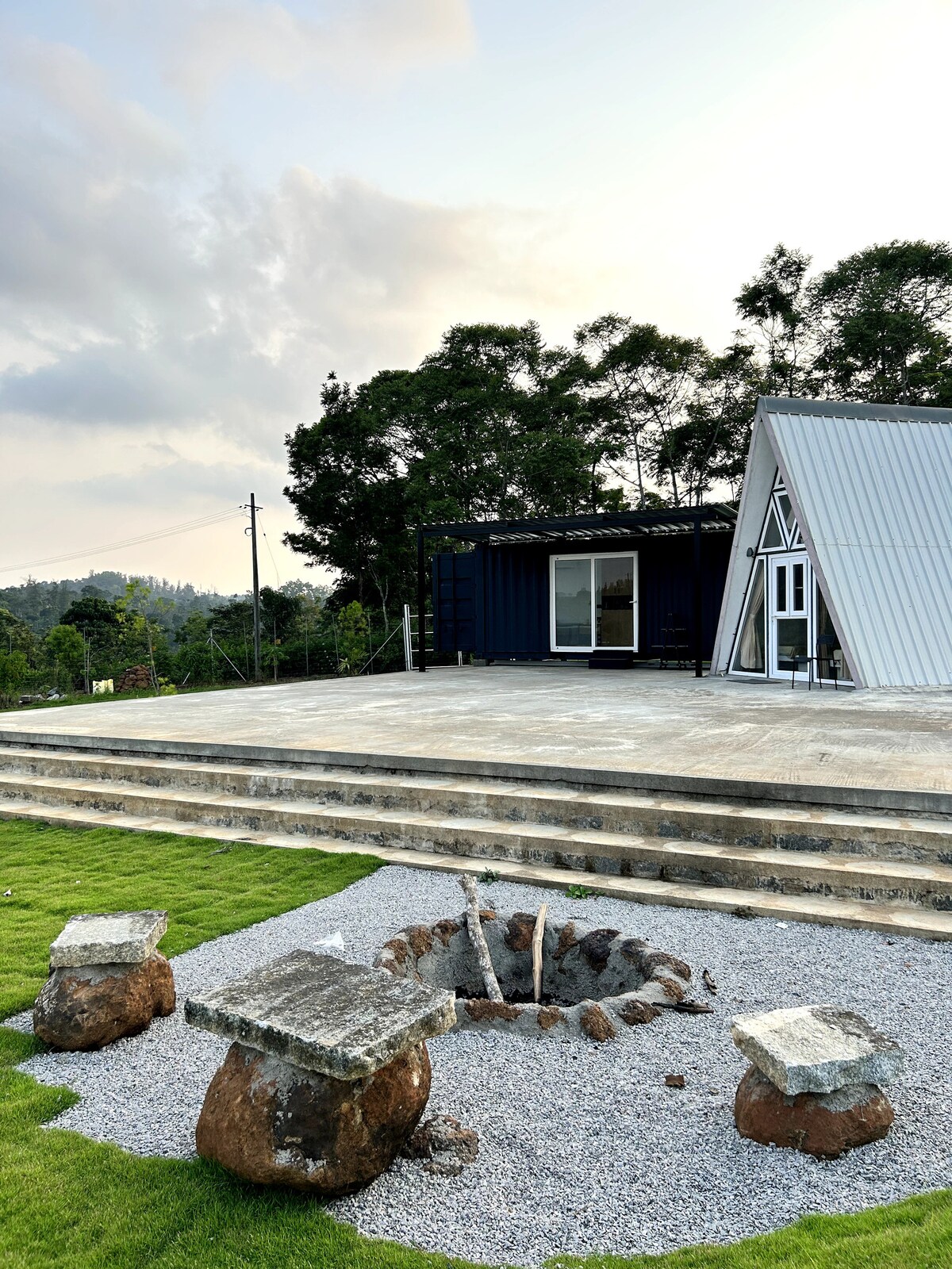 An outdoor area of the retreat features a well-maintained concrete patio, framed by lush greenery. A modern A-frame cabin and a container home stand adjacent, while a circular fire pit made of stones is positioned on gravel, providing a cozy gathering spot.