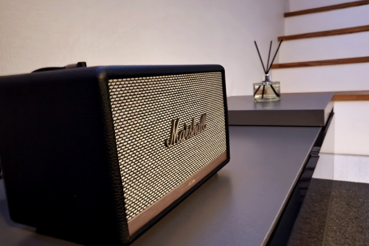 A sleek Marshall speaker is positioned on a dark surface, showcasing its signature black finish and metallic grille. An elegant diffuser with sticks sits nearby, adding a touch of sophistication to the setting. The background features softly-lit steps leading to an upper level.