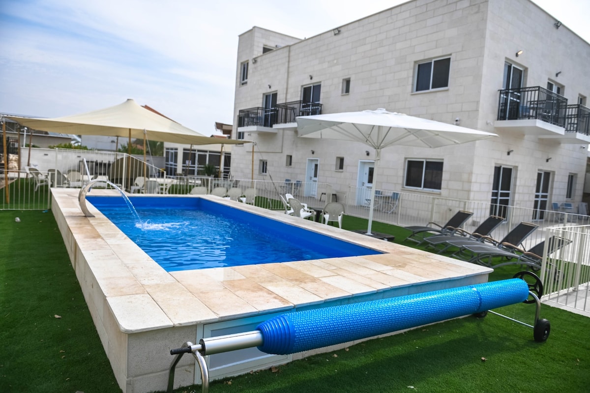 A rectangular swimming pool is centrally featured, surrounded by a paved area and well-maintained green grass. Lounge chairs are arranged alongside the pool, while white umbrellas provide shade. The building in the background features multiple doors and windows, creating a spacious outdoor setting.