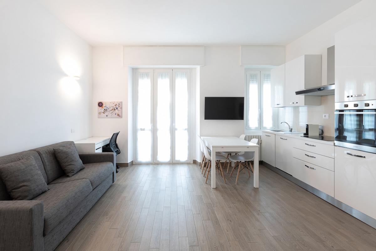 An open living area features a gray sofa, a white dining table with wooden legs, and a sleek kitchen with modern cabinetry. A wall-mounted TV is positioned opposite the sofa. Large windows allow natural light to illuminate the space.