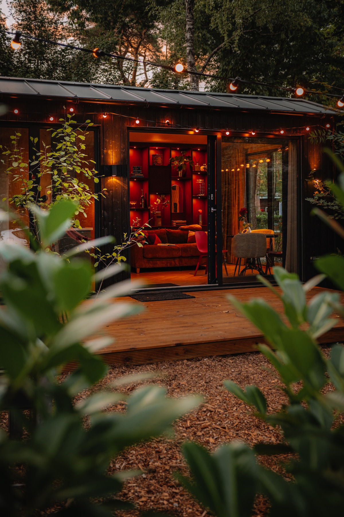 An inviting exterior view of Coco's Buitenhuisje showcases warm lighting from string lights. The entrance opens to a cozy interior filled with rich colors. Surrounding greenery enhances the sense of privacy and natural beauty.