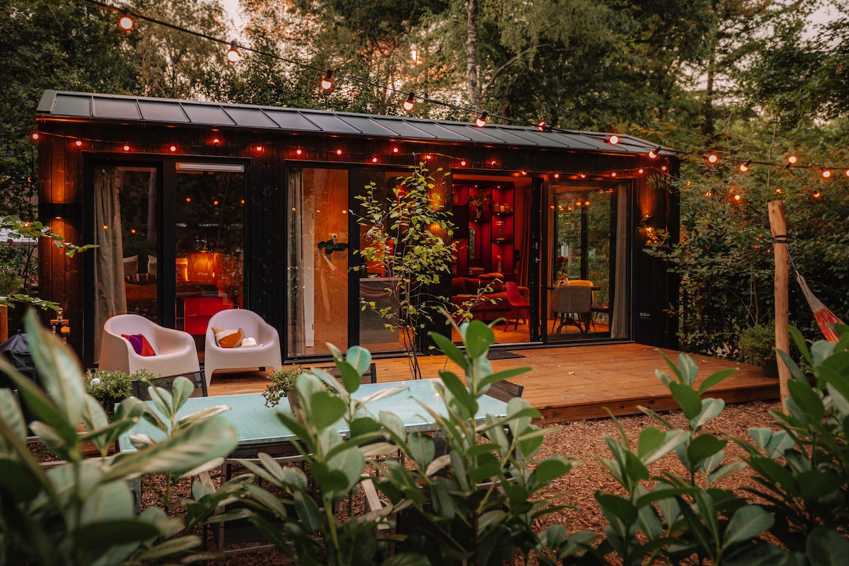 A charming exterior of a tiny house is highlighted by warm string lights above a wooden deck. Two white chairs are positioned beside a table, surrounded by lush greenery. Large glass doors provide a view into a warmly lit interior space.