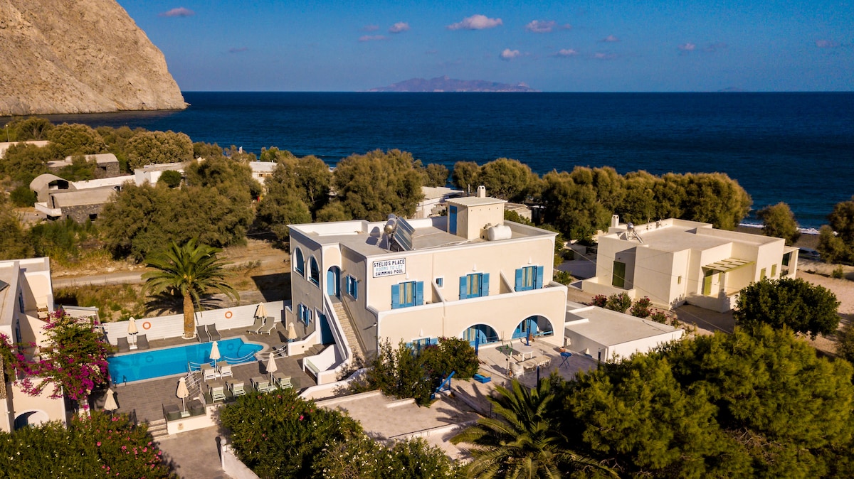 The exterior of Stelios Place is captured, showcasing a light-colored three-story building surrounded by greenery. A pool area is visible in the foreground, accompanied by lounge chairs. The sea can be seen in the background, with rocky cliffs framing the scene and a clear blue sky overhead.