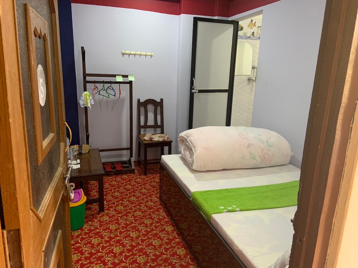 A room featuring a double bed with a light-colored blanket and a bright green decorative throw. A wooden chair and a table are present, alongside a clothing rack with hangers. A door leads to the attached bathroom, while a colorful rug covers the floor.