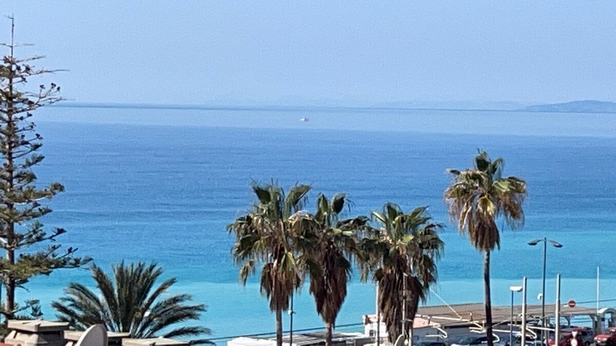 A serene view of the sea showcases varying shades of blue, with palm trees in the foreground. Their fronds sway gently above the coastal landscape, while a small boat is visible on the horizon. The tranquil atmosphere reflects the charm of the Riviera.