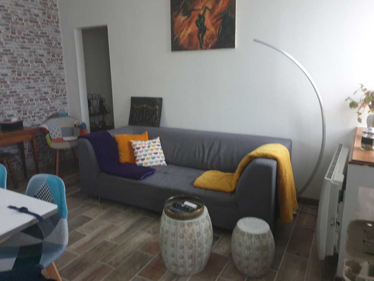 A spacious living area is presented with a gray sofa adorned with colorful throw pillows and a yellow blanket. Two round, textured stools are placed nearby. Wall art adds a touch of charm, while natural light brightens the inviting space.