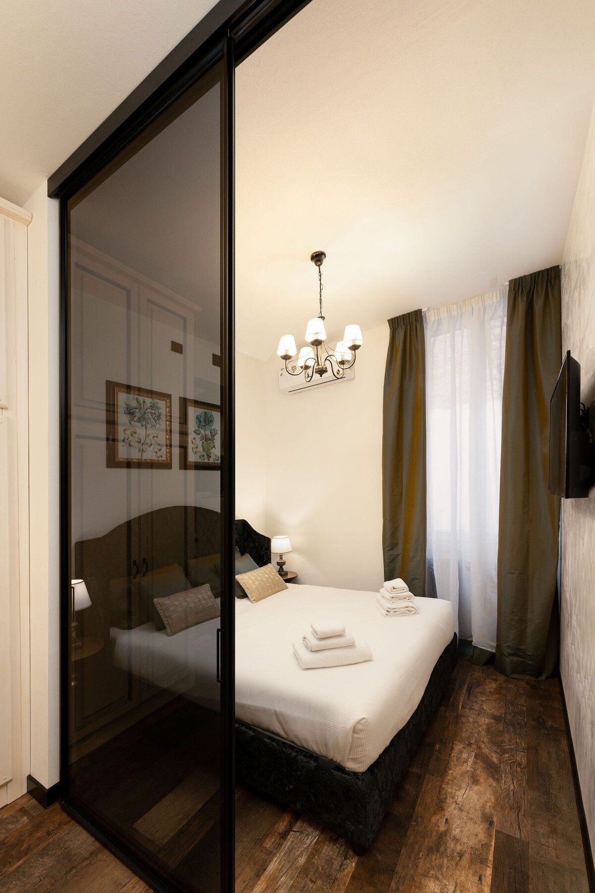 A cozy bedroom is divided from the living area by a sleek smoked glass sliding door. A spacious bed is dressed with fresh linens, complemented by decorative pillows. Warm wood flooring and soft curtains allow natural light to illuminate the space, creating a calm environment.