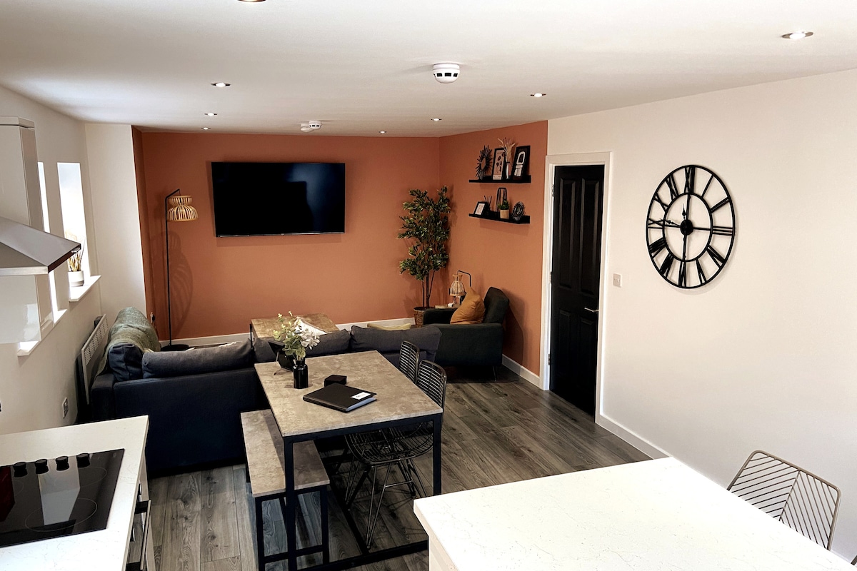 An open living-kitchen area is highlighted by modern decor and a neutral color palette. A spacious sofa is positioned in front of a wall-mounted television. A dining table for four is set on a textured floor, complemented by stylish seating. Artwork and plants enhance the inviting space.