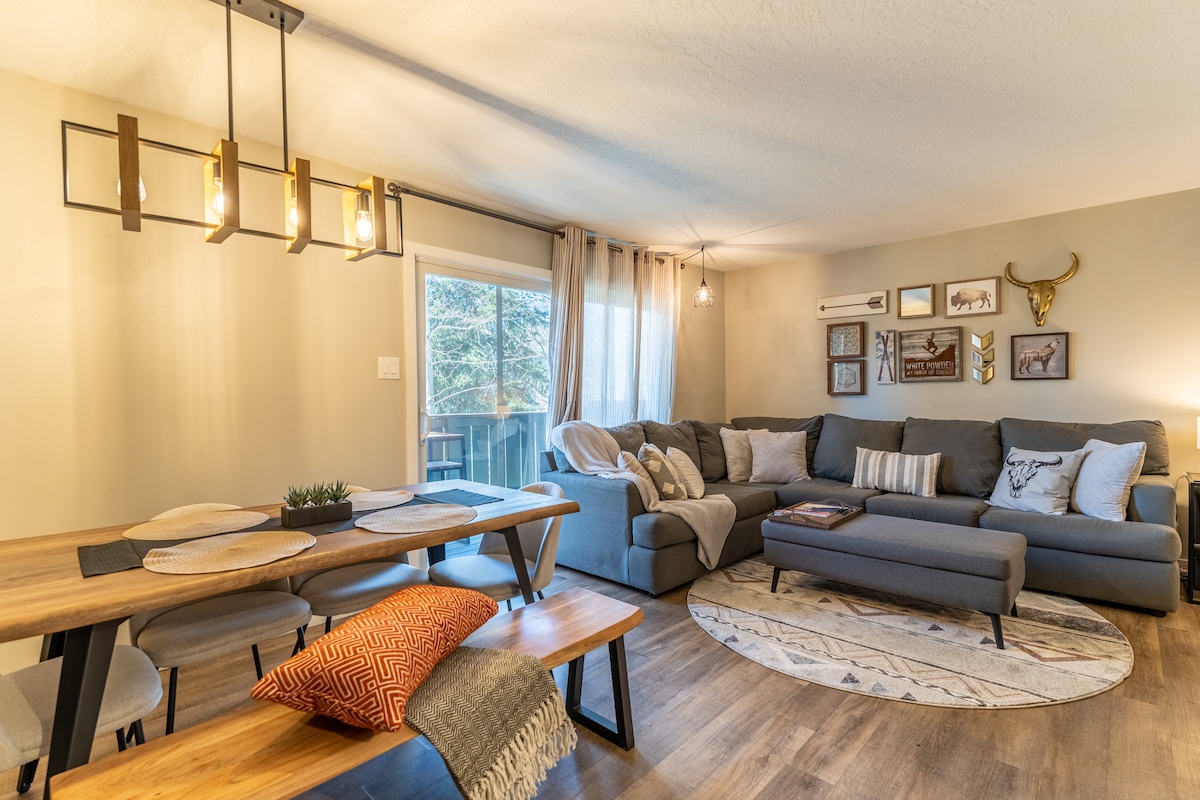 An inviting living area features a spacious gray sectional sofa accented with patterned cushions. A wooden dining table is set with decorative plates and a small potted plant. Natural light filters through large windows, illuminating the cozy atmosphere.