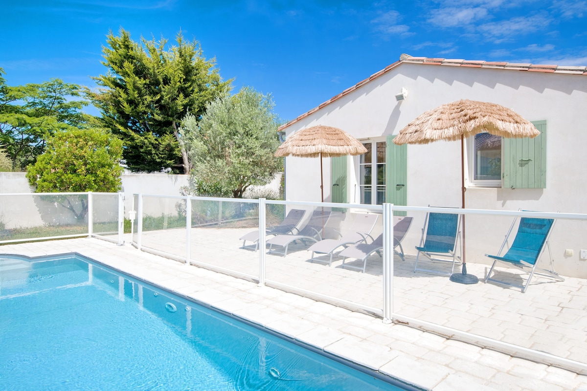 A private pool is surrounded by a protective fence, with two straw umbrellas providing shade over loungers. Green shrubs and trees offer a natural backdrop, while light-colored pavers lead to the whitewashed house featuring green shutters and a window.