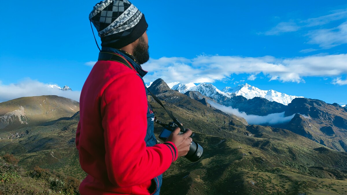 A person stands on a hillside, gazing towards a majestic mountain range topped with snow. The sky is bright blue with scattered clouds, offering a clear view of the peaks. The individual is dressed warmly, wearing a hat and holding a camera.