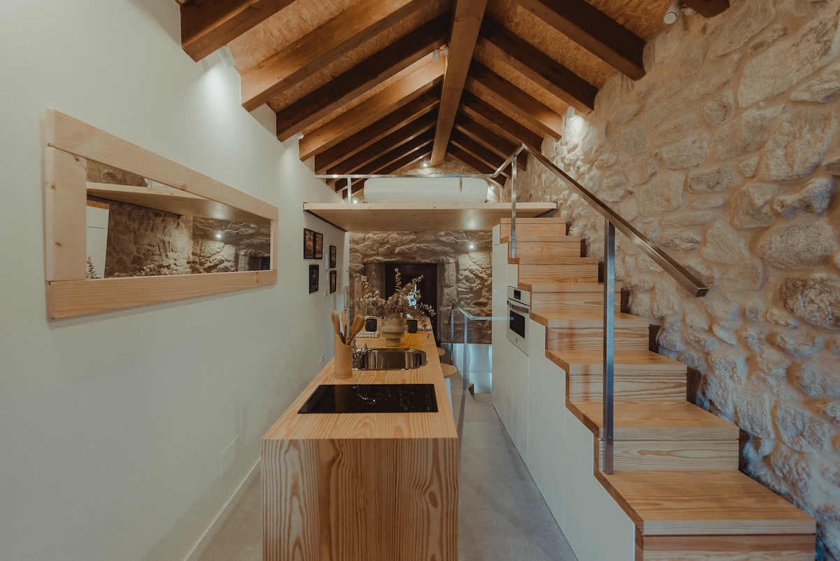 The kitchen area features a modern wooden island with a sink and preparation space. Natural stone walls and wooden beams create a rustic yet contemporary ambiance. A staircase leads to an upper level, while soft lighting enhances the inviting atmosphere.