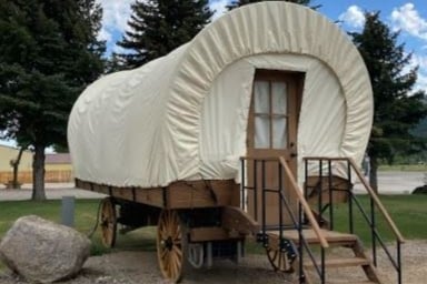A Conestoga wagon is displayed, featuring a fabric canopy and wooden wheels. The entrance is equipped with a wooden door and a set of steps leading up to it. Surrounding greenery and a large stone are visible, set against a blue sky with scattered clouds.