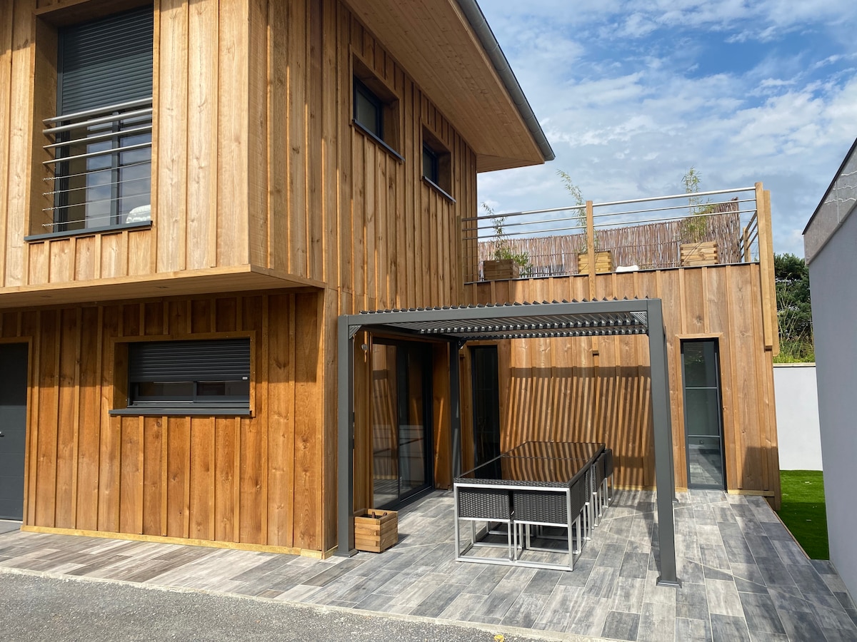 A modern villa exterior is presented, featuring wood-paneled walls and large windows. A covered patio with a long dining table is visible. The space is complemented by a rooftop terrace, with a glimpse of greenery in the background and a clear blue sky above.