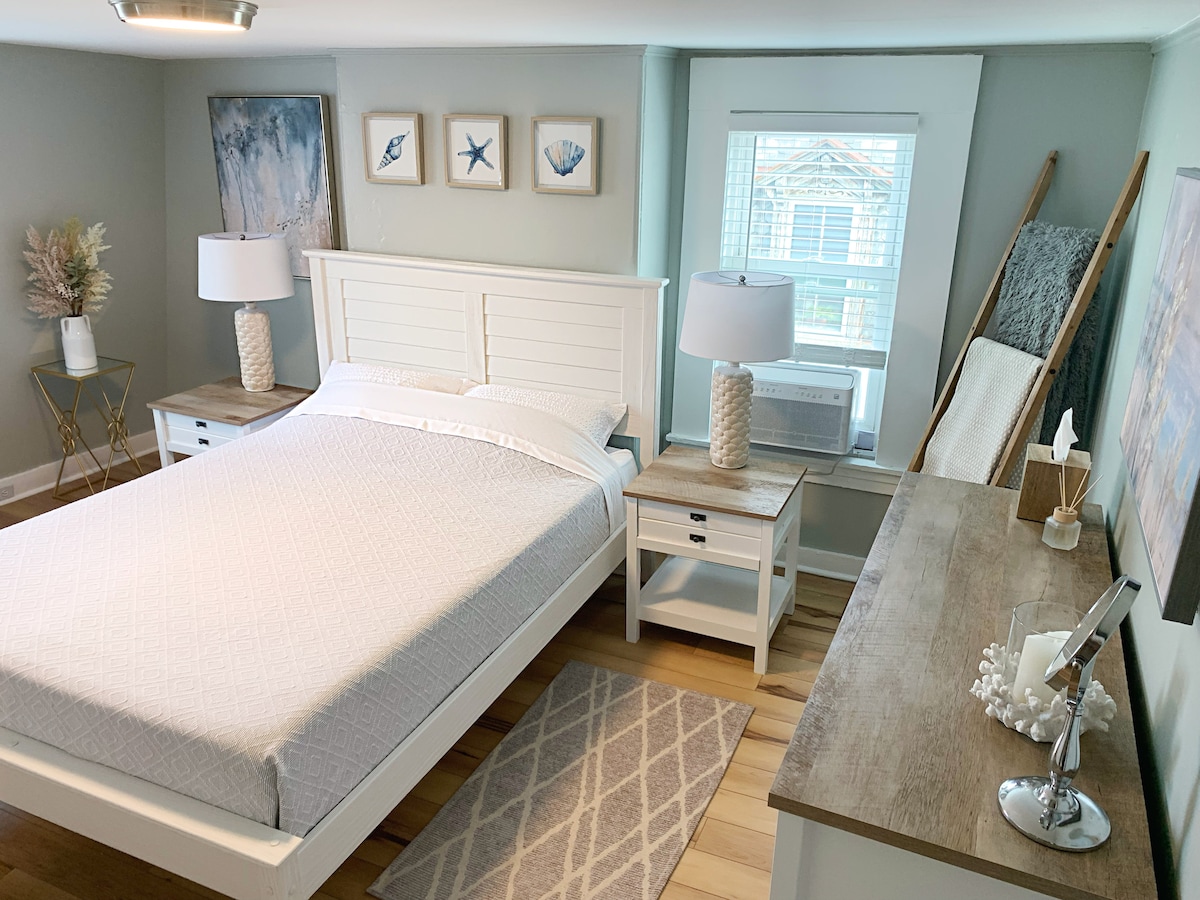 A spacious bedroom features a comfortable queen-sized bed with a light-colored quilt and two bedside tables. Natural light enters through a window adorned with sheer curtains. Decorative elements include framed coastal art and a textured area rug, enhancing the room's relaxing ambiance.