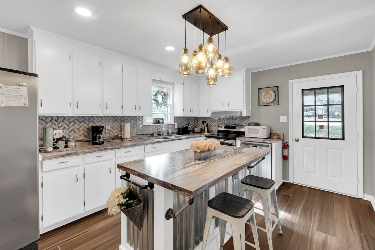 A bright kitchen features white cabinets and a modern design, highlighted by pendant lighting above a central island. A mix of stainless steel appliances and wood elements can be seen, along with a cozy dining area that invites communal meals.