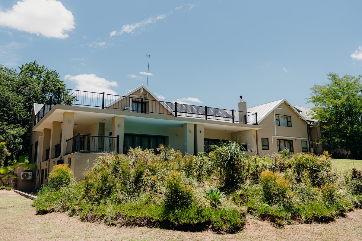 The exterior of a spacious two-story house is framed by lush greenery and landscaped gardens. A balcony extends from the upper level, while solar panels are installed on the roof. The light-colored façade complements the bright sky, providing an inviting appearance.