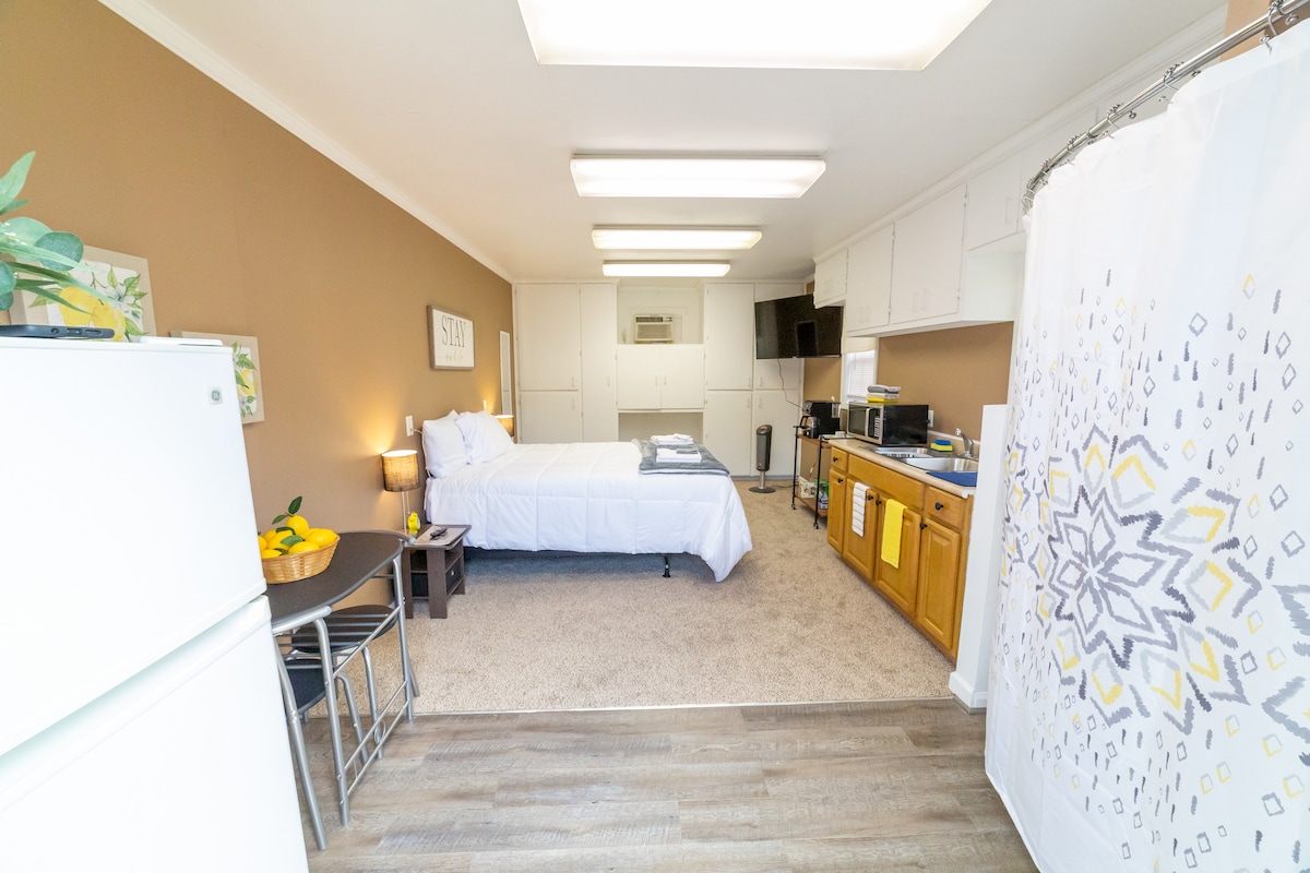 This studio features a spacious sleeping area with a comfortable bed dressed in white linens. A kitchenette is equipped with a refrigerator, microwave, and cabinets. Light-colored walls enhance the open feel, while a decorative shower curtain adds a touch of style.