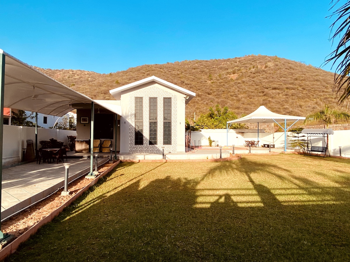 A spacious outdoor area features a well-maintained lawn surrounded by mountains. A modern villa with large windows stands prominently, complemented by shaded seating areas under canopies. The clear sky adds to the peaceful ambiance, creating a perfect setting for relaxation.