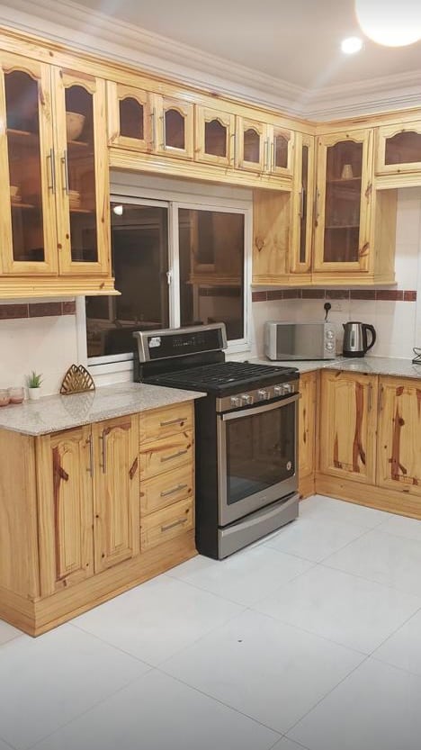 The kitchen features natural wood cabinets with glass-front doors, complemented by a sleek stove and stainless steel microwave. A window allows natural light to illuminate the countertop, which is equipped with essential appliances and decorative accents.