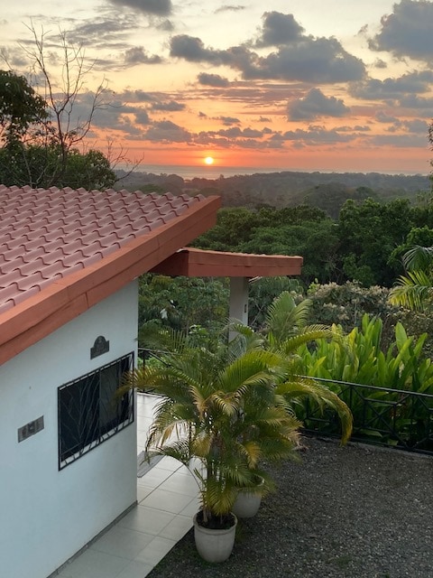 A tranquil sunset is visible beyond lush jungle vegetation, with the sun casting warm hues over the horizon. The exterior of the villa is framed by a terracotta roof and a small potted palm plant, contributing to the serene ambiance of the surrounding landscape.