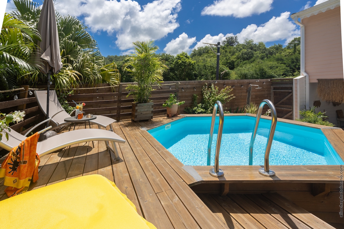 A private outdoor deck is equipped with lounge chairs and a vibrant yellow mattress. A small pool, framed by greenery, is prominently featured, reflecting the blue sky above. The area is designed for relaxation, with a shaded umbrella providing comfortable seating.