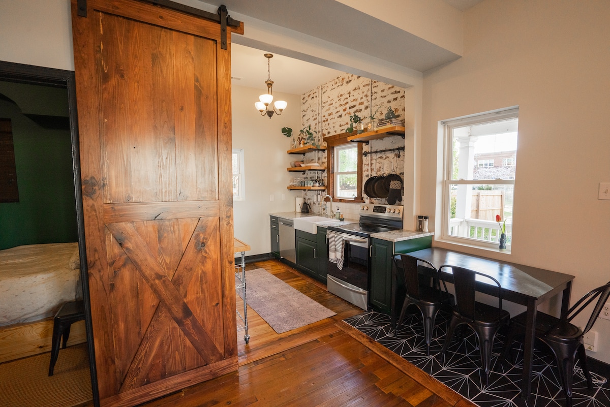 A spacious kitchen area is showcased with a modern stove and white cabinetry. Open shelving holds decorative items and cookware above a dark countertop. A dining table for two is positioned beside a window, allowing natural light to illuminate the space. A rustic sliding barn door adds character.