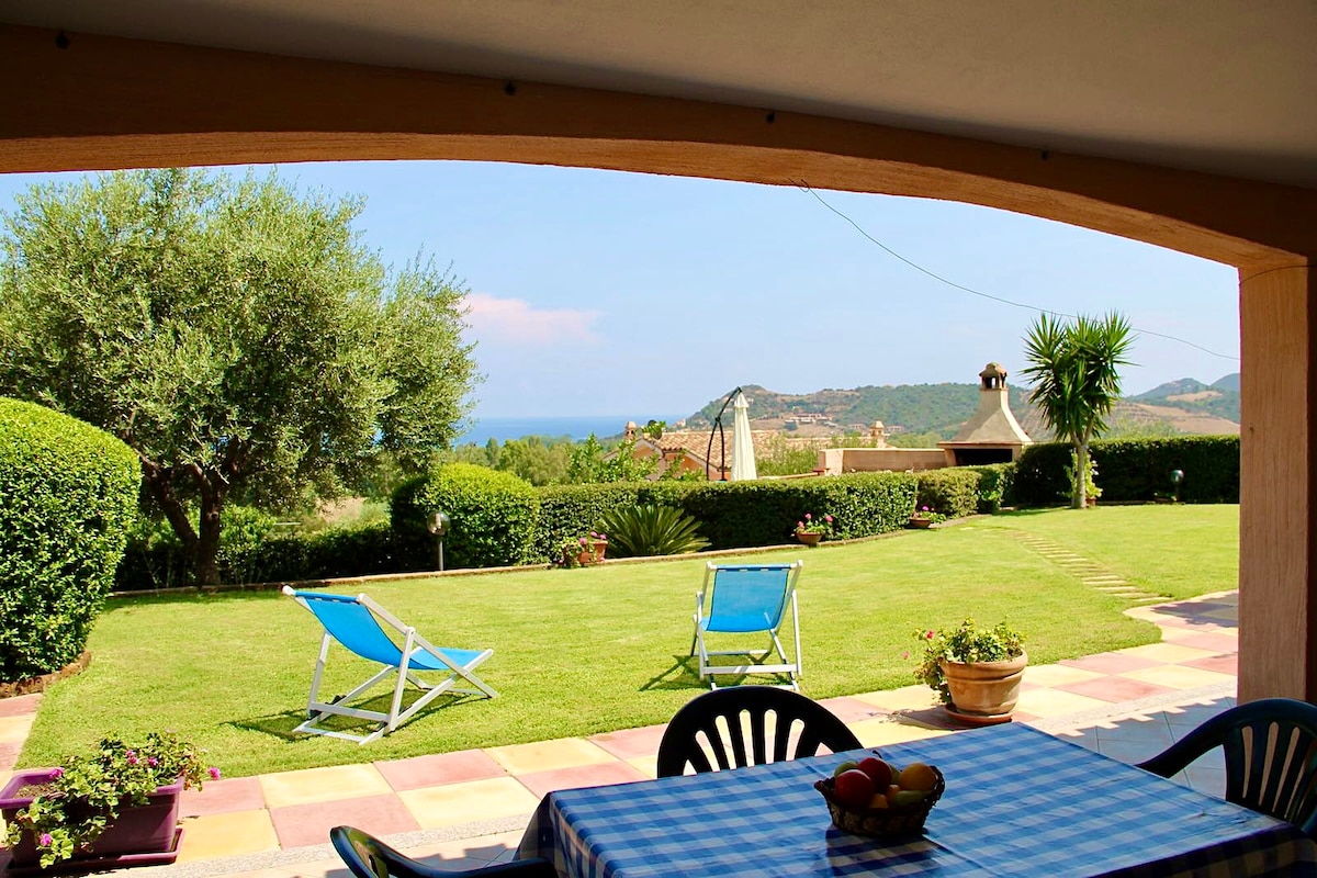 A shaded veranda provides a view of the lush green garden and ocean in the distance. Two lounge chairs are positioned on the lawn, while a dining table set with a blue checkered tablecloth is visible under the awning, complemented by potted plants nearby.
