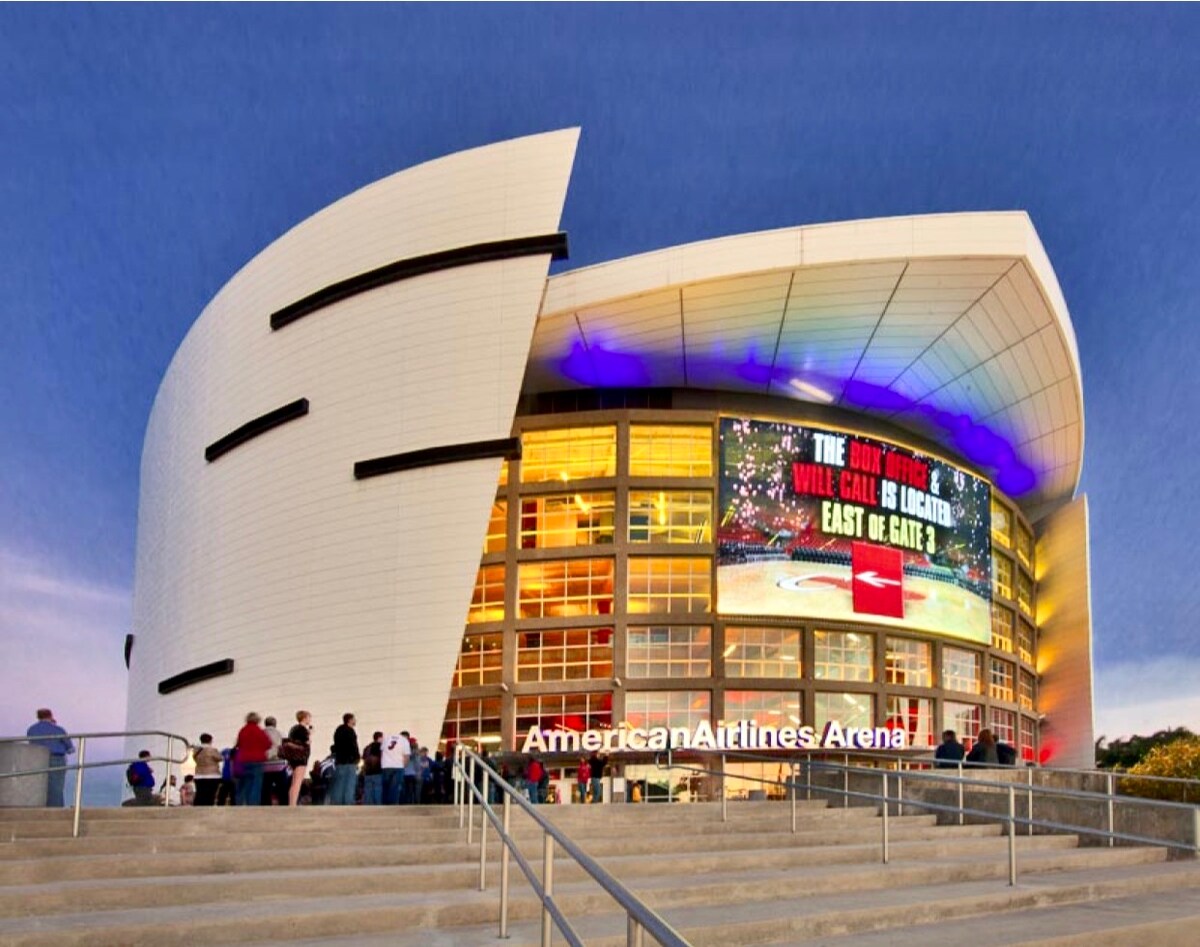 An exterior view of the American Airlines Arena is showcased during twilight. The arena's modern architecture features a curved facade and large glass windows, with vivid lighting highlighting the entrance. A crowd gathers outside, creating a lively atmosphere.