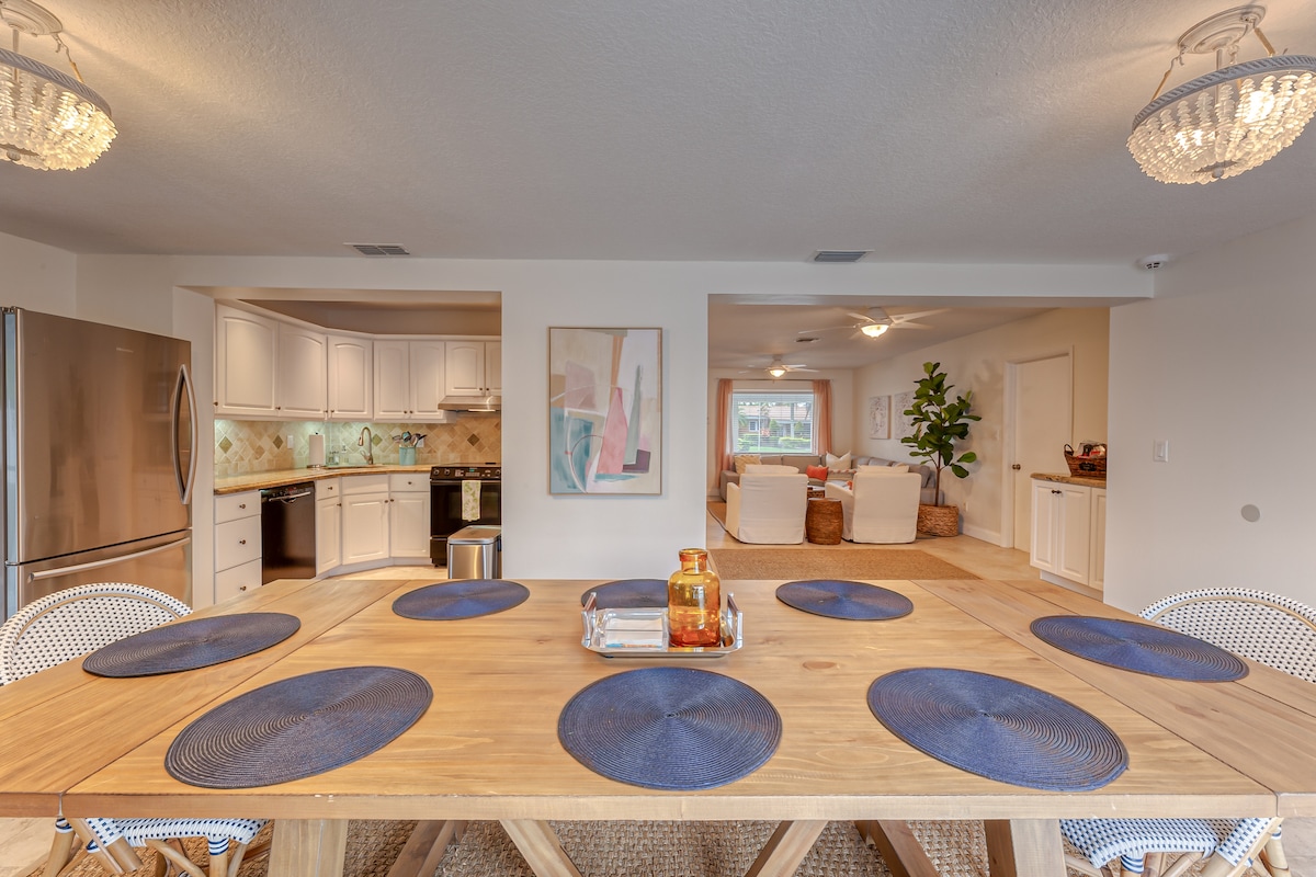 A spacious dining area features a wooden table surrounded by woven chairs. Blue placemats are arranged neatly on the table. A bright kitchen is visible in the background, showcasing white cabinetry and modern appliances. Soft lighting is provided by decorative ceiling fixtures.