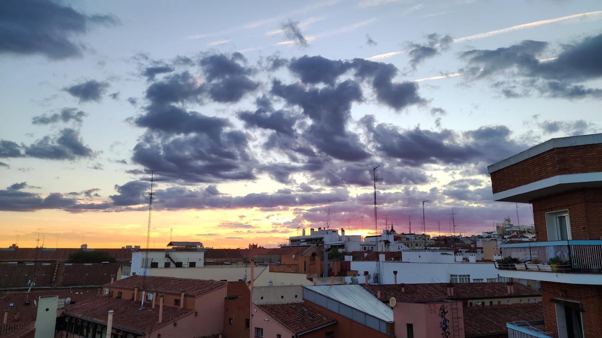 A vibrant sunset fills the sky, casting warm hues over the rooftops of Madrid. Fluffy clouds contrast against the colorful backdrop, while buildings with varied architectural styles are visible below. The image captures a serene moment amidst the bustling cityscape.