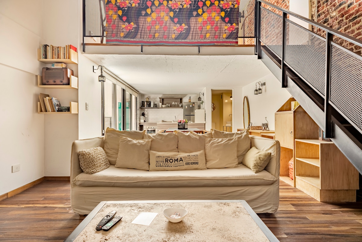 A spacious living room is filled with natural light, featuring a large beige sofa facing a contemporary coffee table. An open staircase leads to a lofted area above. Textured brick walls and wooden flooring add warmth to the space, complemented by decorative shelves and greenery.