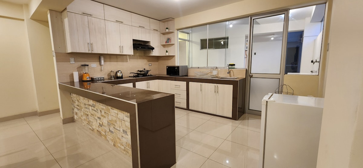 The kitchen features modern cabinetry and a spacious island with stone accents. Natural light fills the area through large windows, illuminating the sleek countertops and appliances, including a refrigerator and oven. The open layout connects seamlessly to the living area.