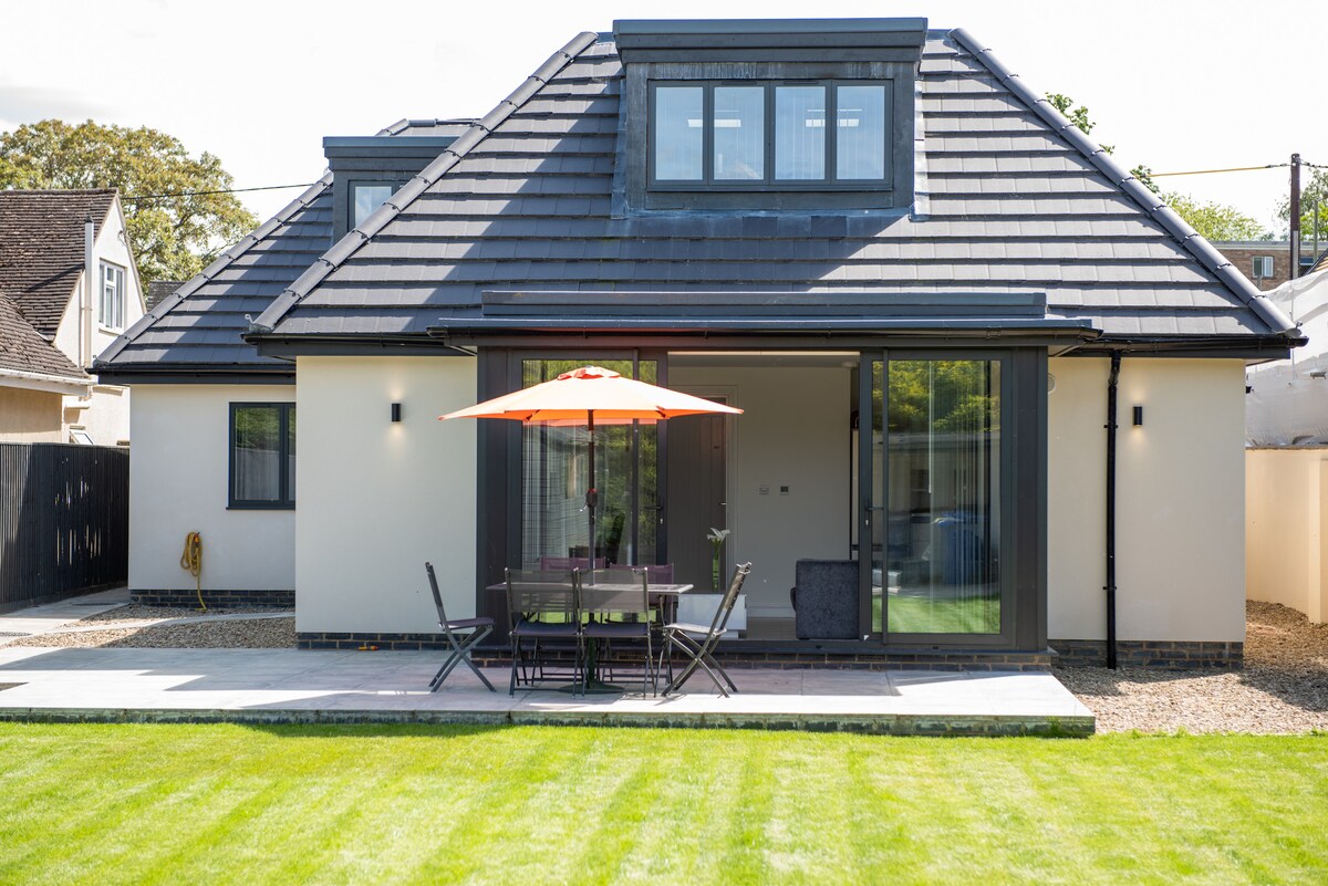 The exterior of the modern apartment is showcased, featuring a sloped roof and large glass doors that open to a patio area. A table with six chairs and a bright orange umbrella provide outdoor seating, while a well-manicured lawn adds to the inviting space.