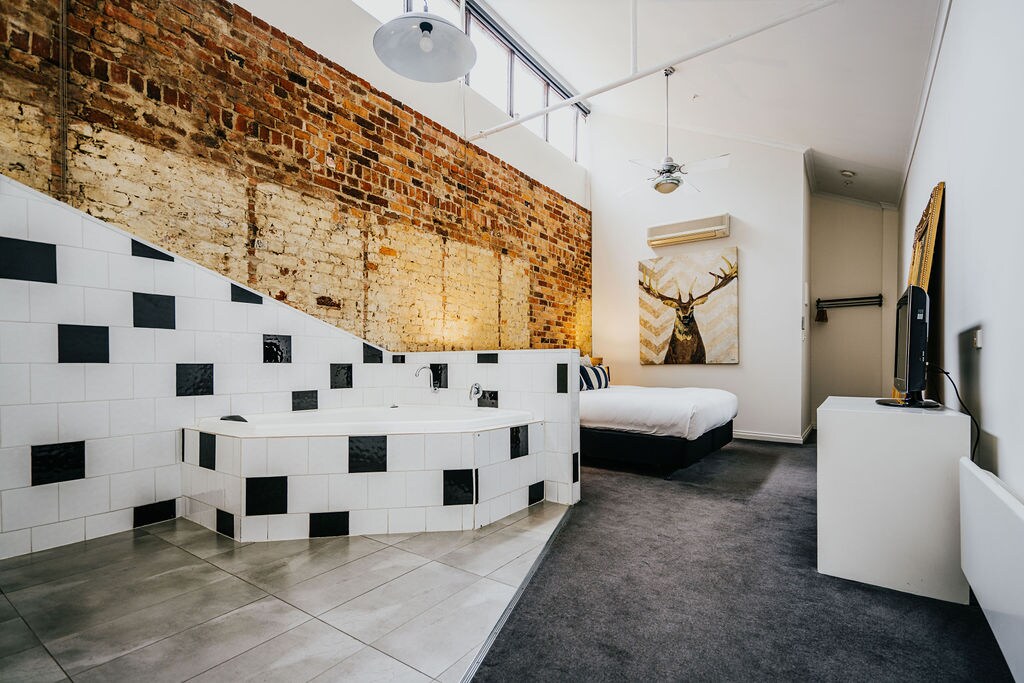 The spacious room features a distinctive exposed brick wall paired with modern white and black tiled accents. A large spa bath is positioned next to the bed, while the cozy sleeping area showcases a king-size bed with a simple, elegant design. Natural light enters through high windows.