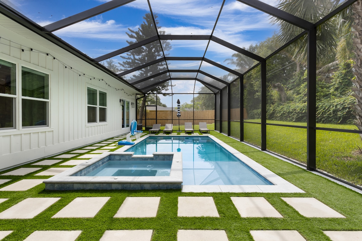 A spacious outdoor area features a screened-in heated saltwater pool and spa, surrounded by a lush green lawn. Comfortable lounge chairs are positioned along the poolside, complemented by decorative pavers. The scene is brightened by natural light filtering through the screen enclosure.