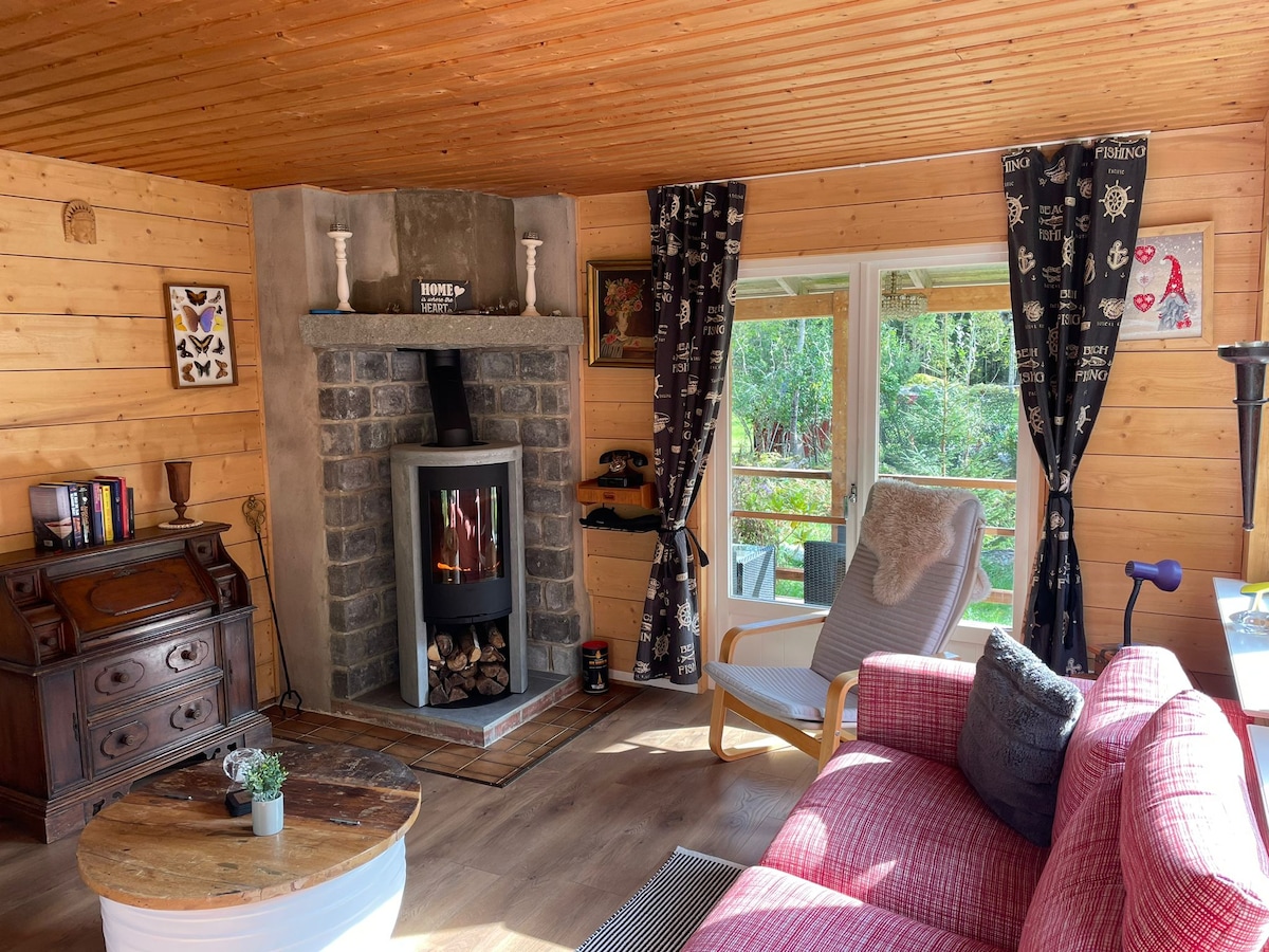 A cozy living area features a stone fireplace at its center, surrounded by natural wood paneling. Comfortable seating is provided by a red sofa and a reclining chair. Natural light enters through large windows, adorned with dark patterned curtains. A small table with a potted plant adds a touch of greenery.