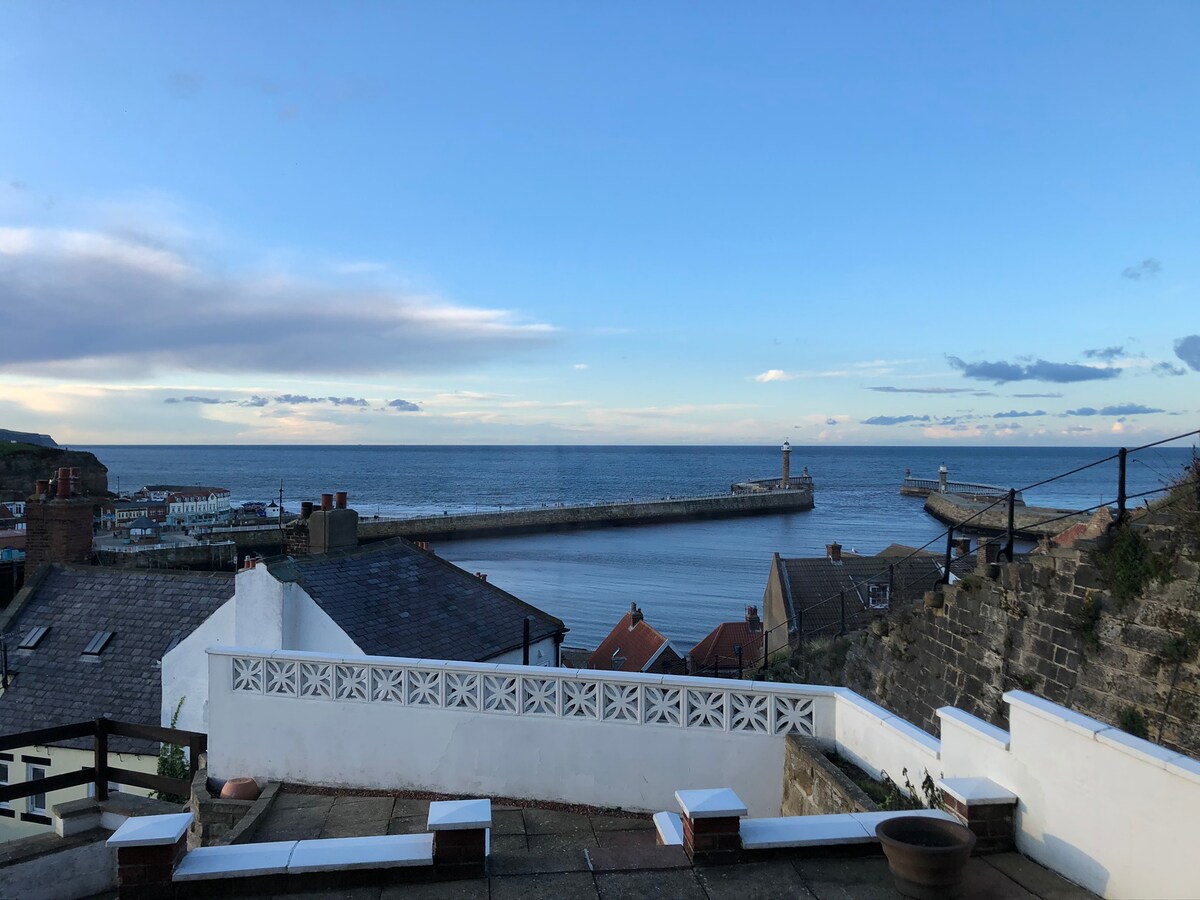 An expansive view of the sea and harbor is showcased from a terraced area, featuring a distant lighthouse and boats along the shoreline. The horizon is painted with soft clouds and a light blue sky, creating a serene backdrop to the coastal landscape.