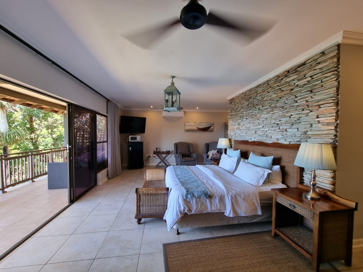 An elegantly designed room features a king-size bed with light bedding, complemented by a natural stone wall. Large glass doors open to a private balcony, while a sitting area includes two chairs and a small table. Soft lighting is provided by a ceiling fan and lantern fixture.